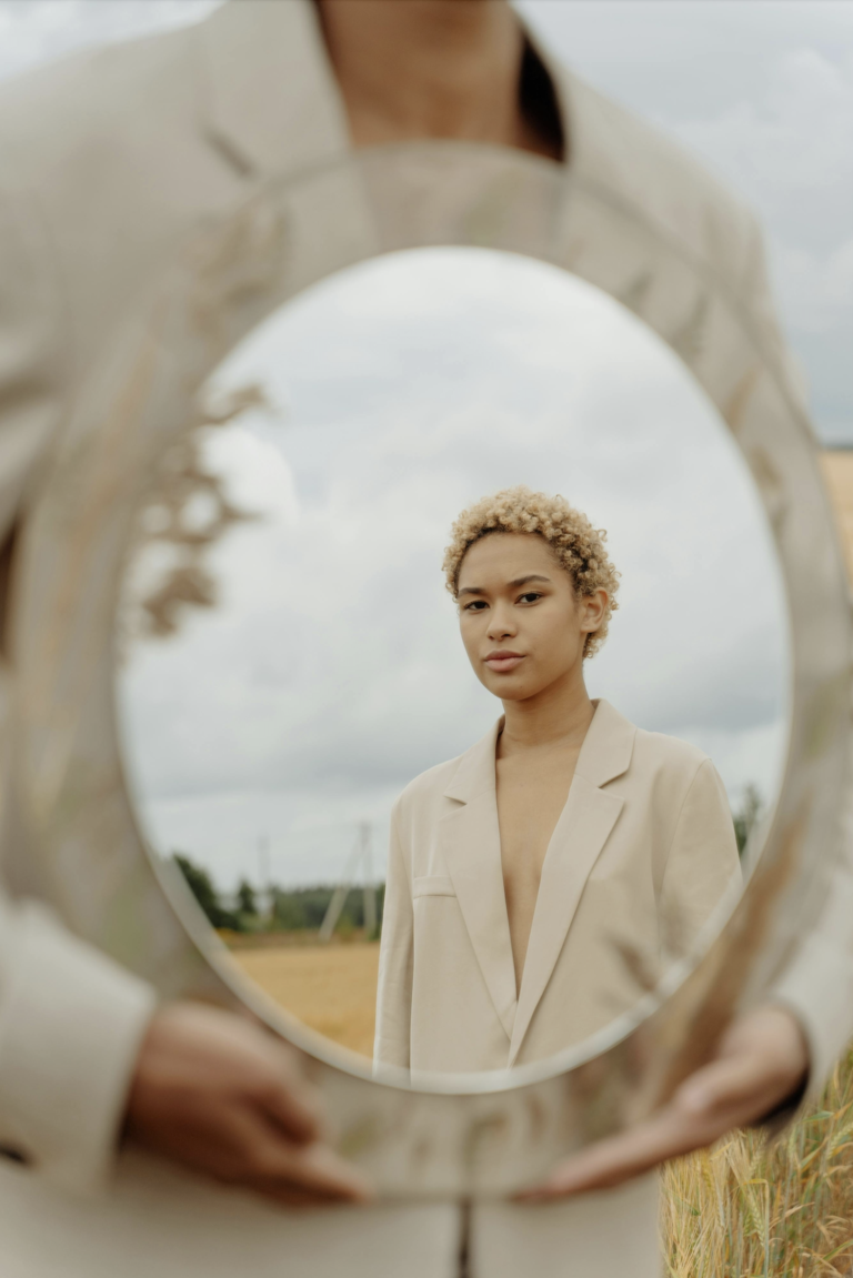 A close up of a person in a field holding an oval mirror that's reflecting another person with the field and sky behind them.